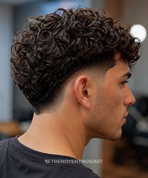 Close-up side view of a young man with defined curly hair and a dramatic mid drop taper fade, showcasing the curved fade blend in soft indoor lighting.