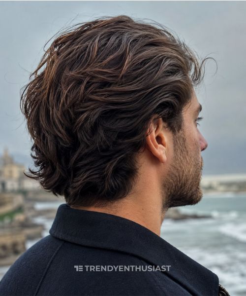 Close-up smartphone photo of a young man with medium-length brown hair featuring a subtle tapered neckline beneath flowing layers, photographed from a rear three-quarter angle against a softly blurred coastal background.