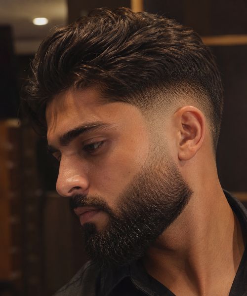 Close-up of a man with a mid taper fade blending seamlessly into a full, well-groomed beard, highlighting the smooth sideburn transition under warm indoor lighting.