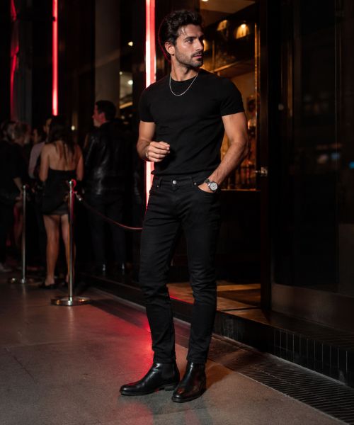 All-black men's club outfit with black T-shirt, slim-fit jeans, and Chelsea boots outside a British nightclub at night.