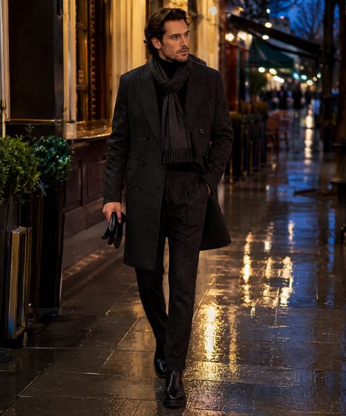Winter men's night out outfit with charcoal overcoat, black turtleneck, scarf, and Chelsea boots on a rain-soaked city street.
